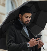 Man looking at his phone holding a black open umbrella protecting him from the rain.