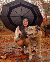 Lady in autumn woods with red-orange leaves, holding an umbrella, taking a photo, petting her dog