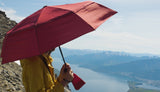 A person in a yellow jacket holds a red umbrella while standing on a mountain, overlooking a serene lake and distant snowy mountains.
